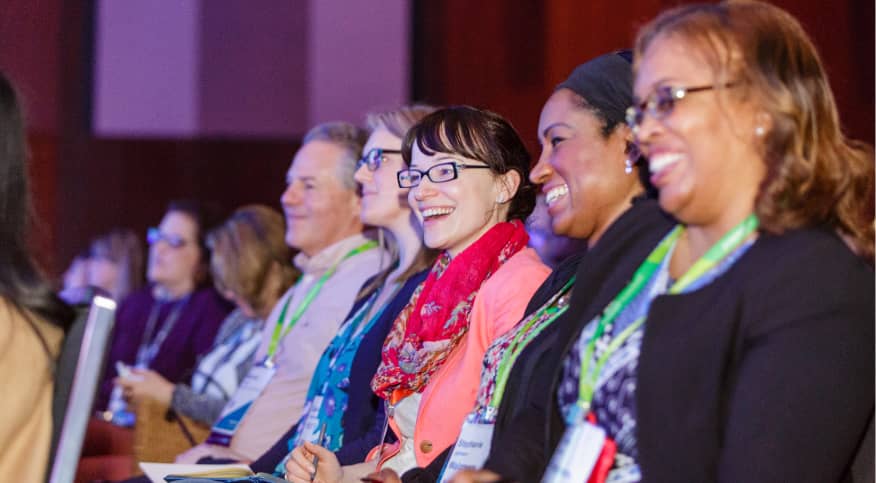 A group of smiling attendees at an event, engaged and enjoying the moment. They are seated in rows, with a diverse mix of individuals. Some are wearing name tags, and several have colorful attire, including a woman in a bright red scarf. The setting has a formal atmosphere, likely at a conference or seminar, enhanced by soft lighting.