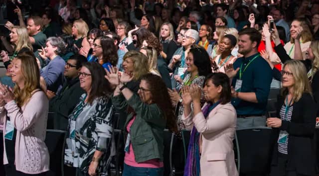 A large, engaged audience is seen at a conference, with attendees smiling, clapping, and cheering. Many people are wearing conference badges, and the atmosphere is vibrant and enthusiastic. The audience is predominantly composed of individuals of diverse backgrounds, all focused on the event happening before them.