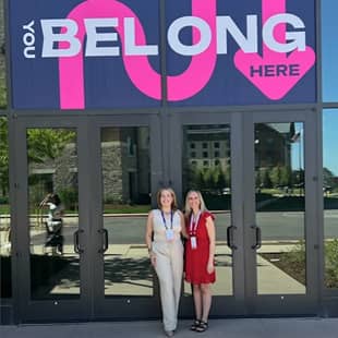 Two women stand in front of a large glass entrance featuring a sign that reads "YOU BELONG HERE" with an arrow pointing down. The first woman, dressed in a light-colored outfit, is on the left, while the second woman, in a red dress, is on the right. The background shows a landscaped area outside the building.