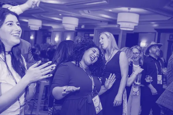 A lively gathering with attendees enjoying music and engaging with each other. A woman in a black dress and curly hair is singing and having fun in the foreground, while others in the background also appear joyful. Various people are cheering and clapping, creating an energetic atmosphere. The setting features well-lit chandeliers and a stylish interior, indicating a celebratory occasion.