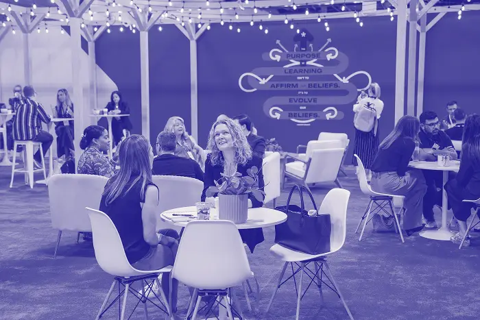 A lively gathering at a conference with attendees engaged in conversation at white circular tables. The atmosphere is enhanced by string lights above. In the background, there are graphics suggesting ideas related to purpose and beliefs, highlighting keywords such as "Learning," "Affirm," and "Evolve."
