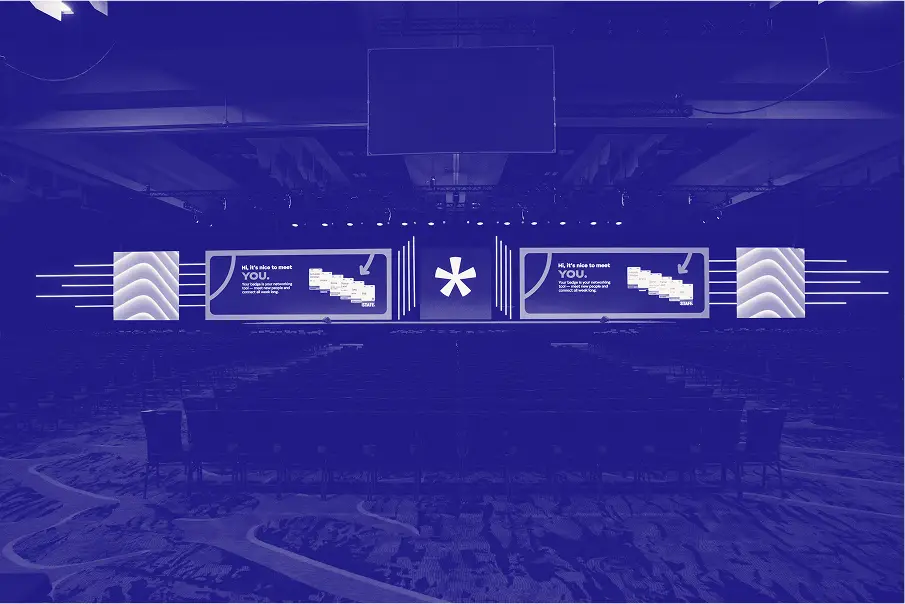 A convention stage setup featuring a large backdrop with the message 'Hi, it's nice to meet YOU' alongside graphical elements and an asterisk symbol. The auditorium is mostly empty with rows of chairs in the foreground, indicating it is set up for an event or presentation. The overall color tone is deep blue, creating a cohesive visual theme.