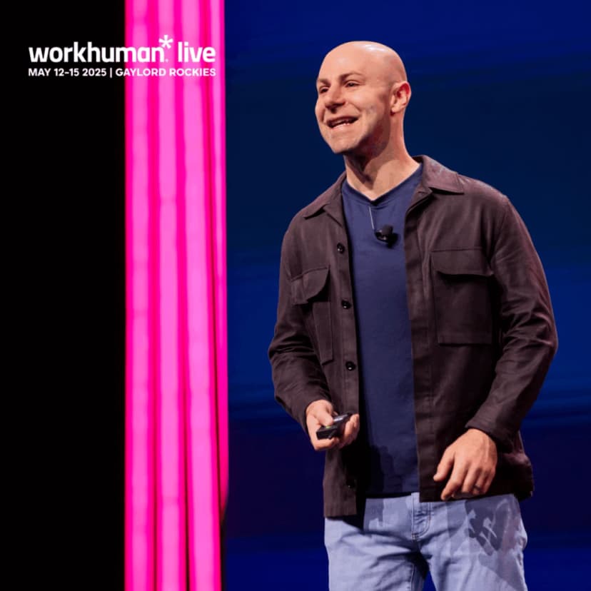 A man smiling while speaking on stage at a Workhuman Live event, with vibrant pink lights in the background. 