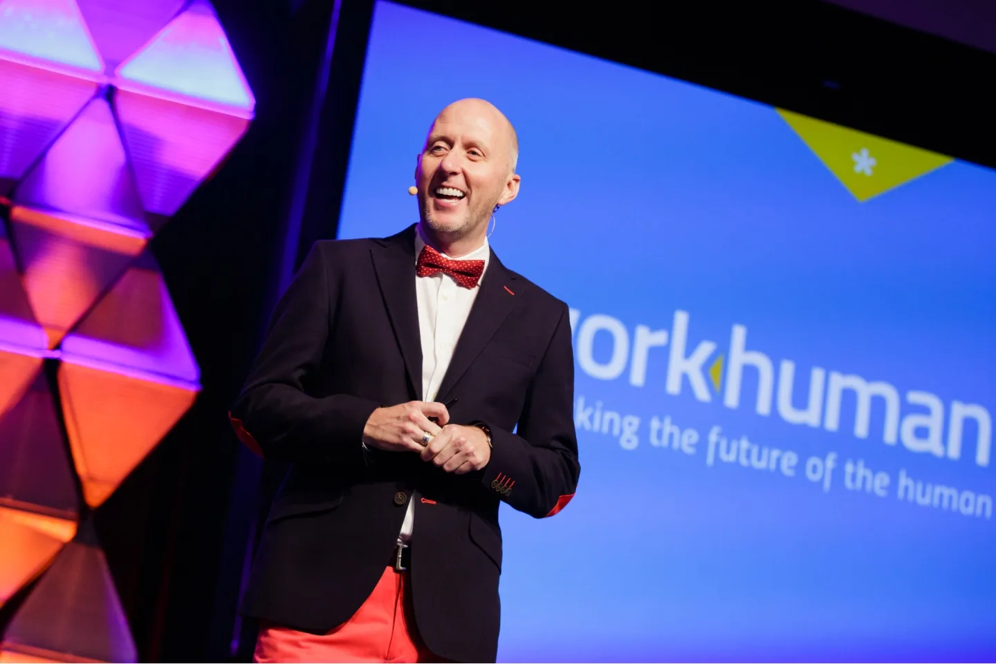 A presenter smiling while holding a microphone stands on stage, wearing a black suit jacket and a red bow tie, paired with bright red pants. Behind him is a blue banner displaying the Workhuman logo and the words "making the future of the human." Colorful geometric shapes in purple and orange are visible in the background, creating an engaging atmosphere.