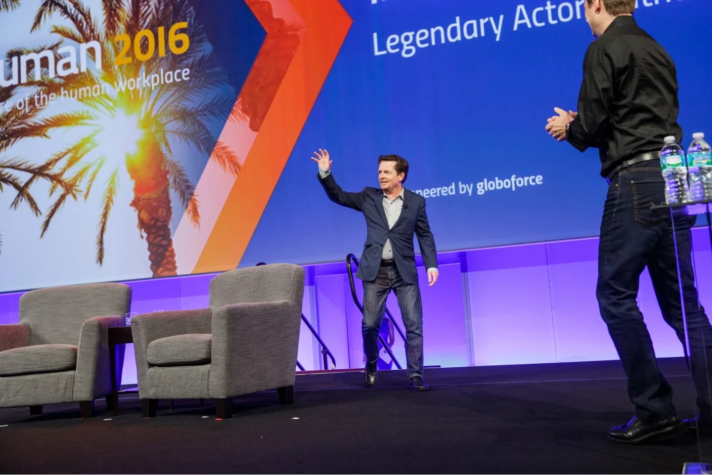 A man gestures energetically while standing on a stage, welcoming the audience at the Workhuman 2016 event. Behind him is a large screen displaying the event's title, "Workhuman 2016," along with the phrase "Legendary Actor of the Human Workplace." Two gray armchairs are positioned on the stage, and several bottles of water are visible on a nearby table, indicating a prepared setting for speakers or guests. The backdrop features a bright, sunny image with palm leaves, adding a vibrant touch to the scene.
