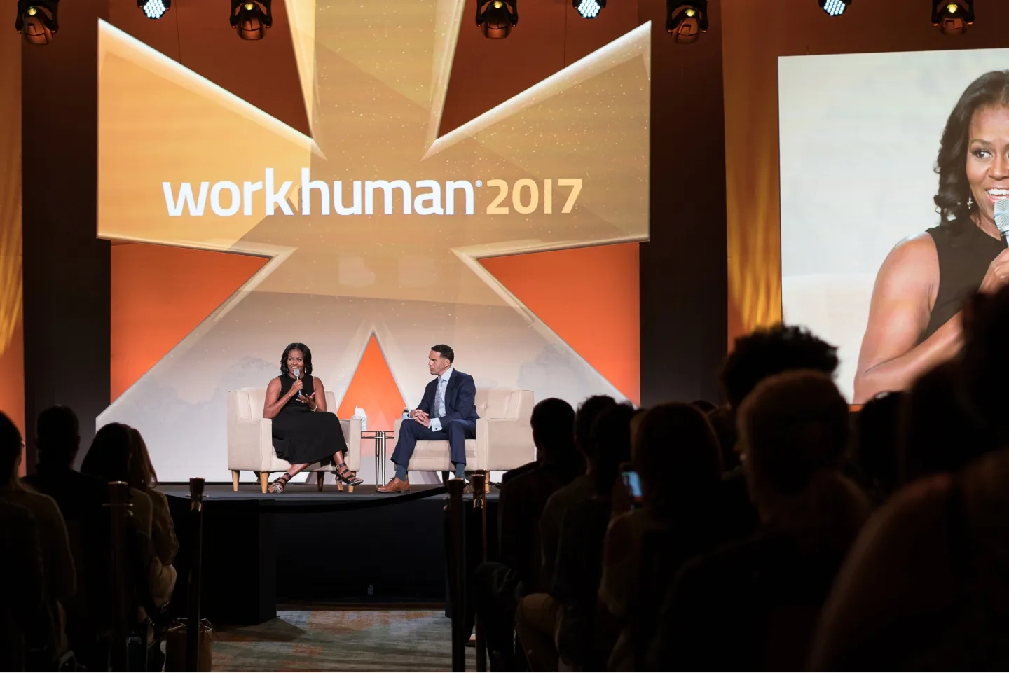 A panel discussion at the Workhuman 2017 conference featuring a speaker on stage. The speaker is seated in a chair with a microphone, while another person, also seated nearby, listens attentively. The background displays the "workhuman 2017" logo alongside an orange and gold star motif. An audience can be seen in the foreground, watching the presentation.