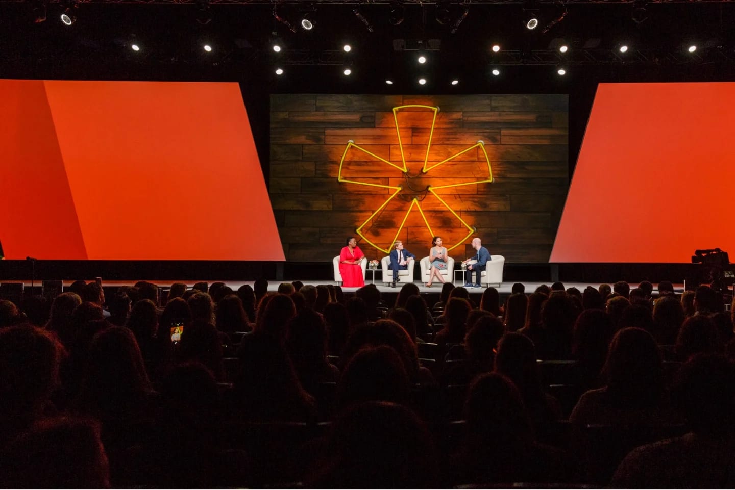 A panel discussion taking place on stage at a Workhuman event. Four speakers are seated in chairs, with three on the right side, while one speaker is on the left in a red dress. The background features a large orange and wood decorative element. An audience is visible in front of them, with many people facing the stage.