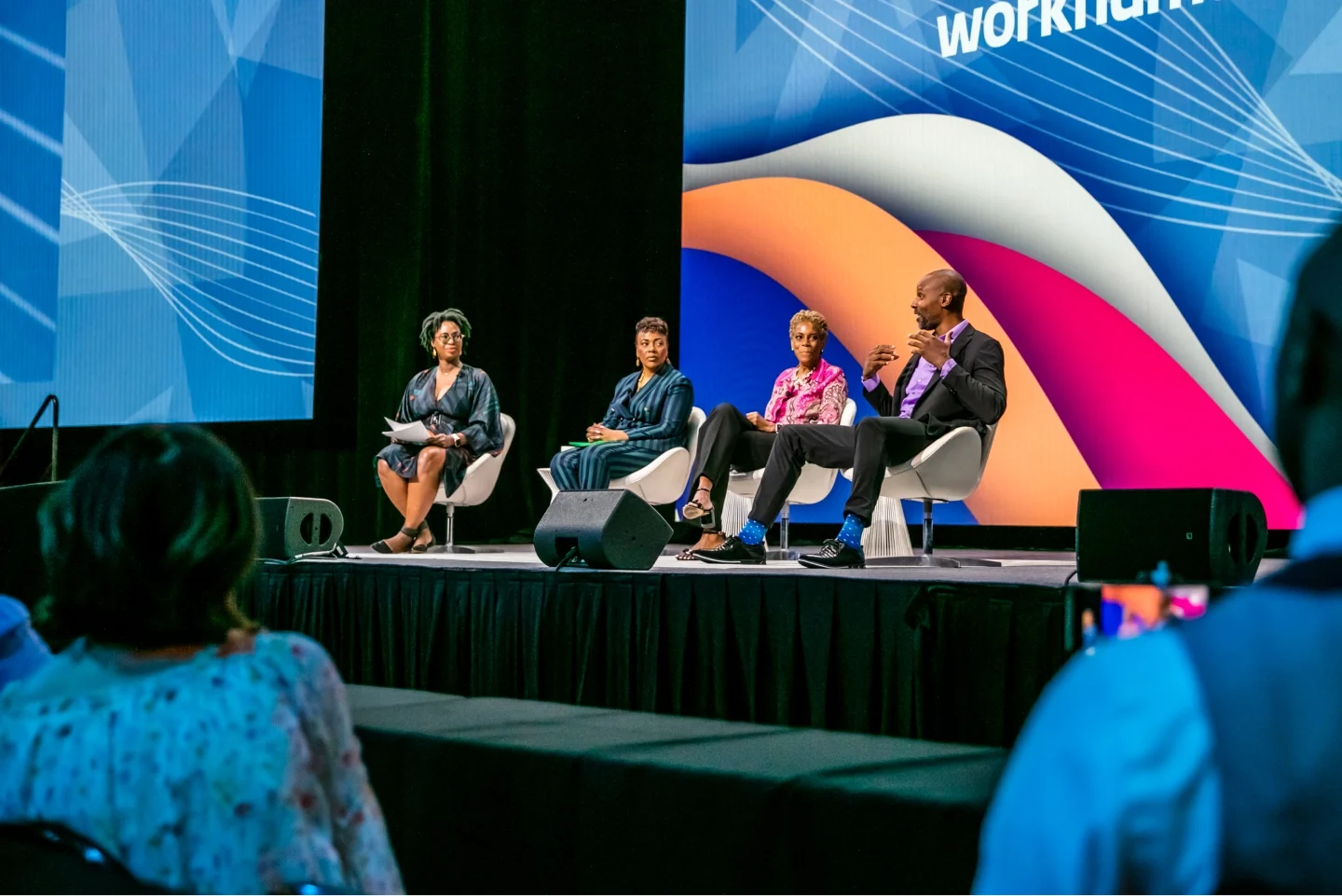 A panel discussion taking place at a Workhuman event, featuring four speakers on stage. The speakers, including a woman on the left holding a microphone and wearing glasses, are engaged in conversation. The backdrop displays colorful graphics related to Workhuman, and the audience is visible in the foreground, attentively listening to the discussion.