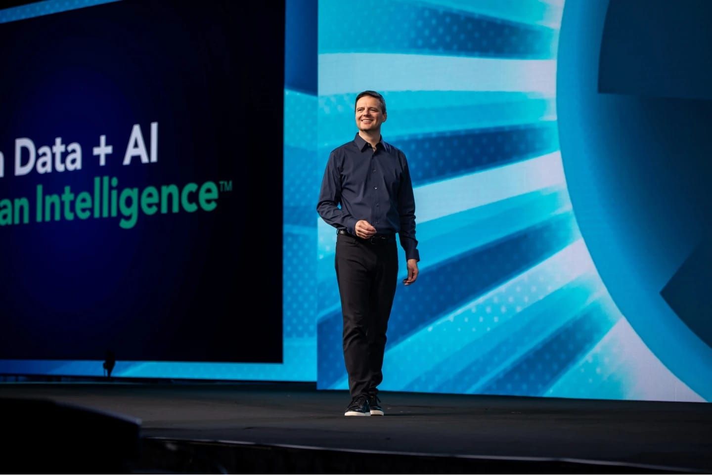 A man in a dark shirt stands on a stage while smiling, with a backdrop displaying the text "Human Data + AI" and "Human Intelligence™" in large, prominent letters. The stage is illuminated with blue and teal lighting, creating a vibrant atmosphere for a presentation.