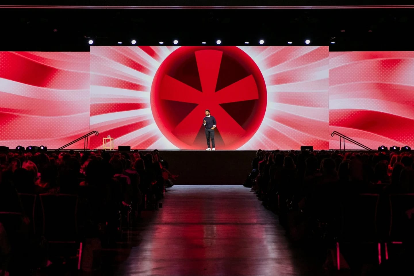 A speaker stands on stage in front of a large screen displaying a vibrant red and white design with a prominent circular element, while an audience sits in rows facing the stage. The setting appears to be an event or conference, and the atmosphere is energetic.