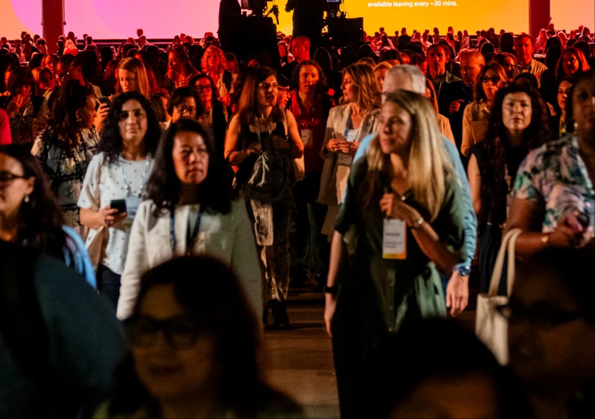 A large group of people is gathered in a brightly lit venue, with a colorful background. Many attendees are walking, some are checking their phones, and several individuals have name badges visible on their clothing.