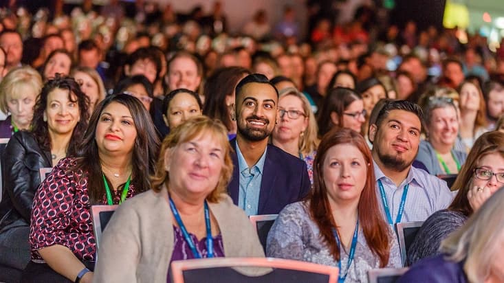 A large audience of diverse individuals is seated in a well-lit venue, with many people smiling and engaging with the event. Several attendees are wearing name lanyards, and the atmosphere appears lively and attentive.