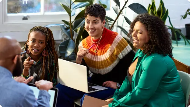A group of three people in a casual meeting, sitting together with laptops in front of them. One person, wearing a colorful knitted sweater, is animatedly speaking and gesturing. They are surrounded by plants in a bright, modern workspace. The atmosphere appears engaging and collaborative, with all participants actively involved in the conversation.