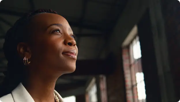 A woman with braided hair is looking upward with a gentle smile on her face, exuding a sense of hope or contemplation. The background features industrial-style windows that let in natural light, creating a warm ambiance. She is wearing a stylish white blazer, contributing to a professional appearance.
