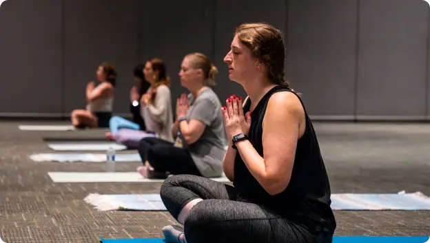 A group of women sitting on yoga mats in a spacious indoor setting, practicing meditation. They are arranged in a row, with their hands in a prayer position close to their chest, indicating a serene atmosphere. The background appears neutral, focusing attention on the participants engaged in mindfulness.