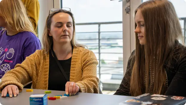 A group of people engaged in an activity around a table with various colorful items, likely part of a workshop or team-building exercise. Two women are prominently featured: one with glasses and a light brown cardigan focused on arranging small pieces, while the other with long, straight hair observes. In the background, other participants are present, and a bright view is seen through large windows. A box labeled with colorful pieces is also visible on the table.