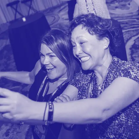 Two women laughing and taking a selfie together, both wearing lanyards, with a blurred indoor setting featuring round tables in the background. Their expressions convey joy and camaraderie as they enjoy their moment.