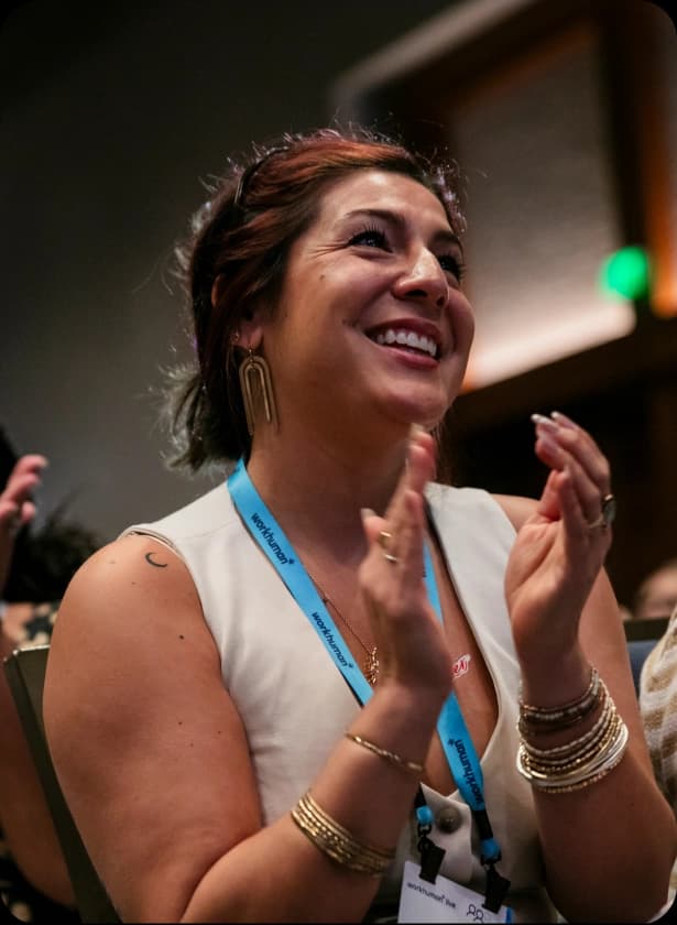 A woman with shoulder-length hair, wearing a white sleeveless top and multiple bracelets, is smiling and clapping her hands in a seated position. She has a blue lanyard around her neck and a crescent moon tattoo on her upper arm, with a softly lit background.