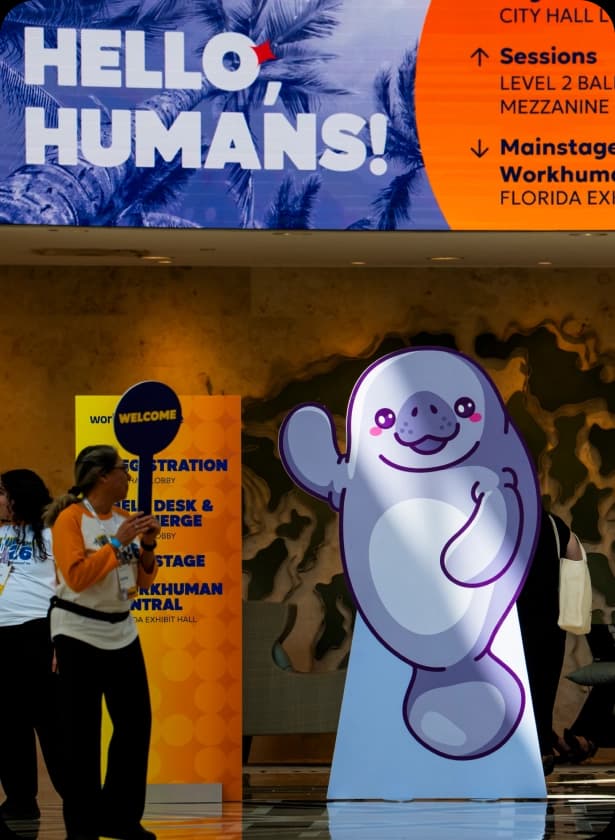 A person in a white shirt with orange sleeves stands near a large, colorful welcome sign in a convention center. The sign displays the text "HELLO, HUMANS!" alongside directional information, while a whimsical, purple manatee figure is positioned prominently nearby.