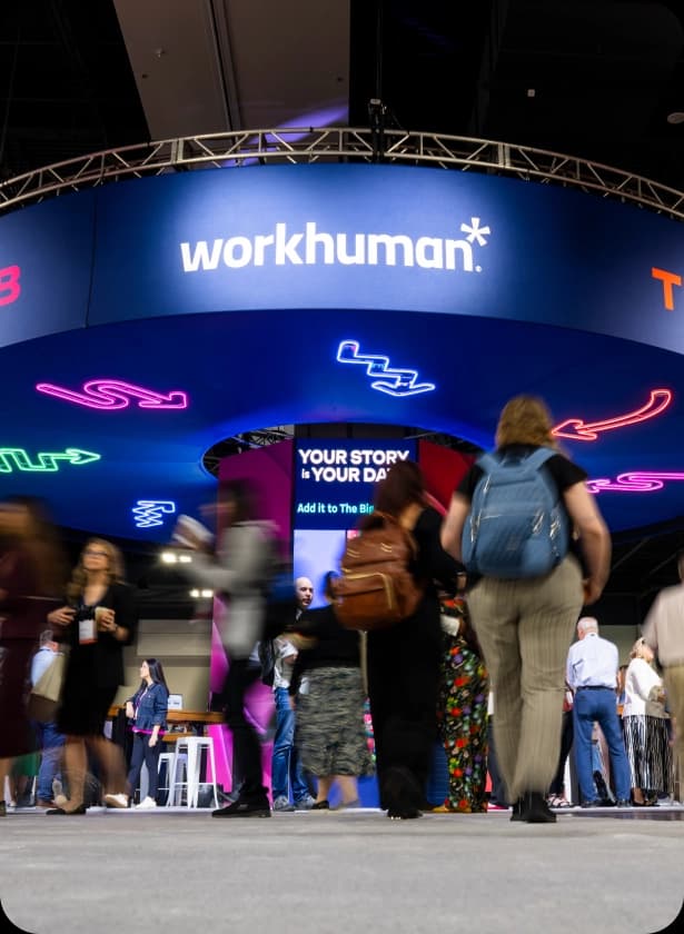 A large exhibition booth with a circular structure featuring the text "workhuman" prominently displayed at the top. Surrounding the booth are colorful neon arrows and the text "YOUR STORY is YOUR DAY" with the phrase "Add it to The Big" beneath it. Attendees wearing various outfits are walking around the booth area.