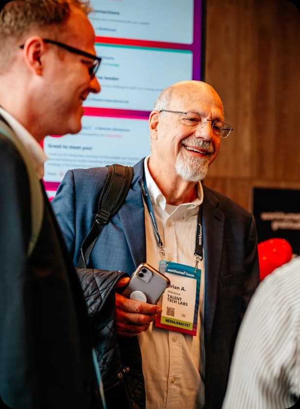 A man with a beard and glasses is smiling while holding a smartphone and standing next to another man in a suit. He is wearing a blazer over a white shirt and has a name tag that reads "Brian A. - Talent Tech Labs." In the background, there are colorful digital displays and a blurred crowd.