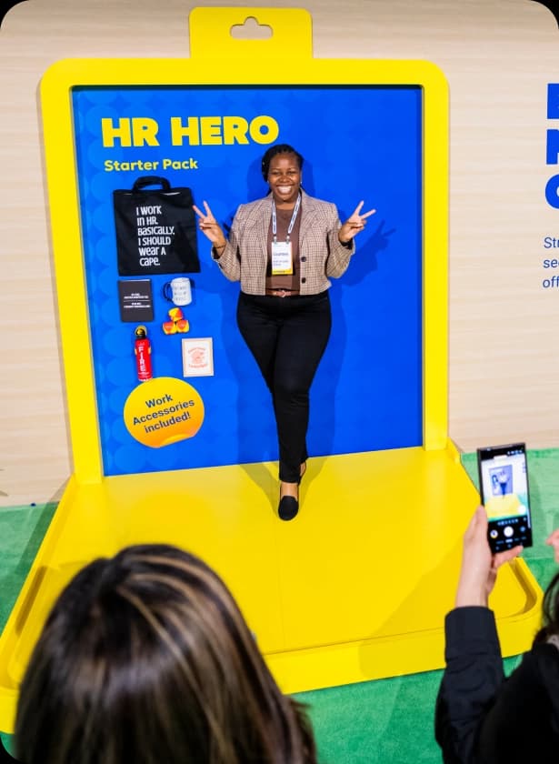 A person stands in front of a colorful backdrop designed like a product display, holding up two fingers in a peace sign. The backdrop features the text "HR HERO Starter Pack" along with various items including a tote bag, a mug, and a water bottle, accompanied by the phrase "Work Accessories included!" Another individual is seen in the foreground, taking a photo with a smartphone.