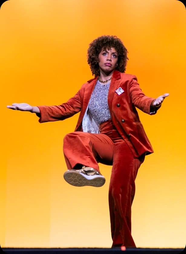 A person with curly hair is wearing a bright orange suit and a shimmering top, standing on one leg with arms outstretched against a solid yellow backdrop. The individual is posing dynamically on stage.