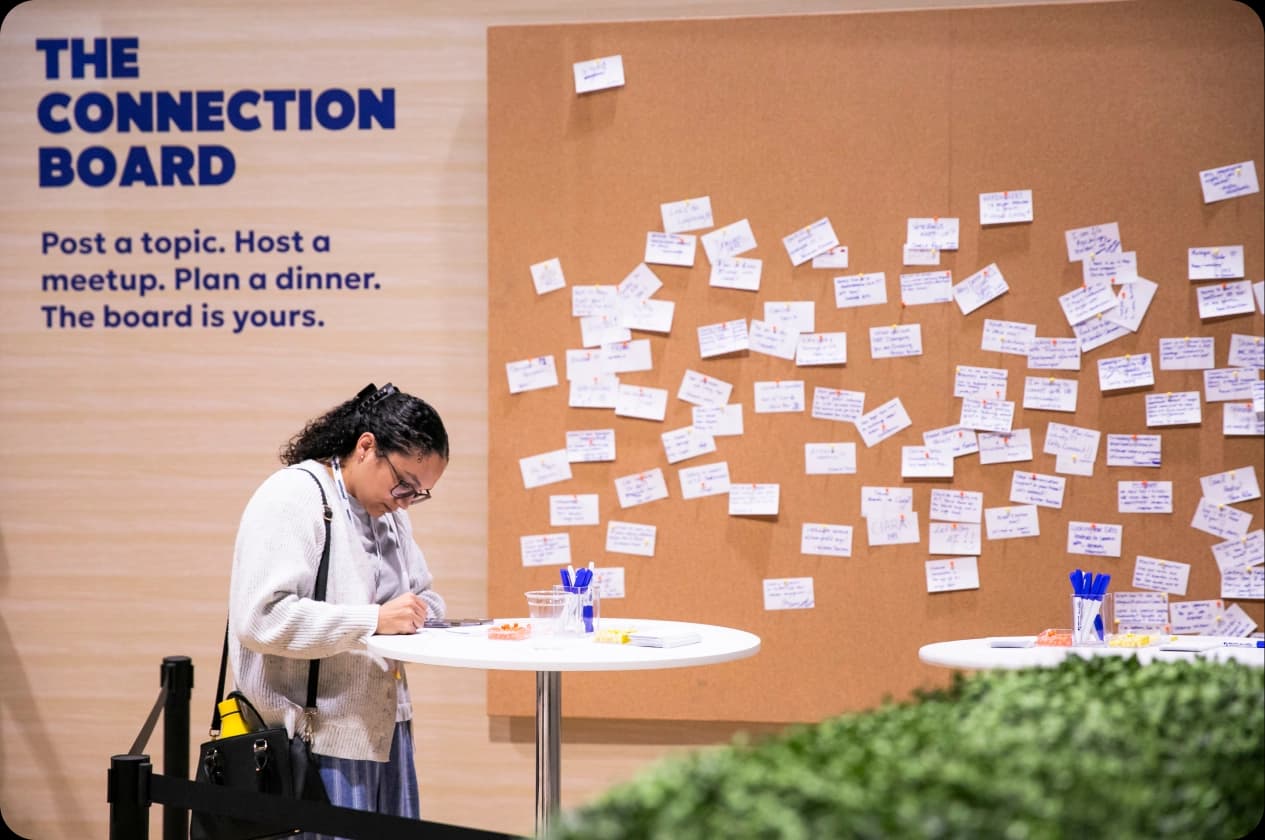 A woman with curly hair is writing on a notebook at a white table in front of a corkboard filled with notes. The corkboard displays multiple pieces of paper with handwritten messages, while a sign above reads, "THE CONNECTION BOARD" followed by instructions to post topics, host meetups, and plan dinners.