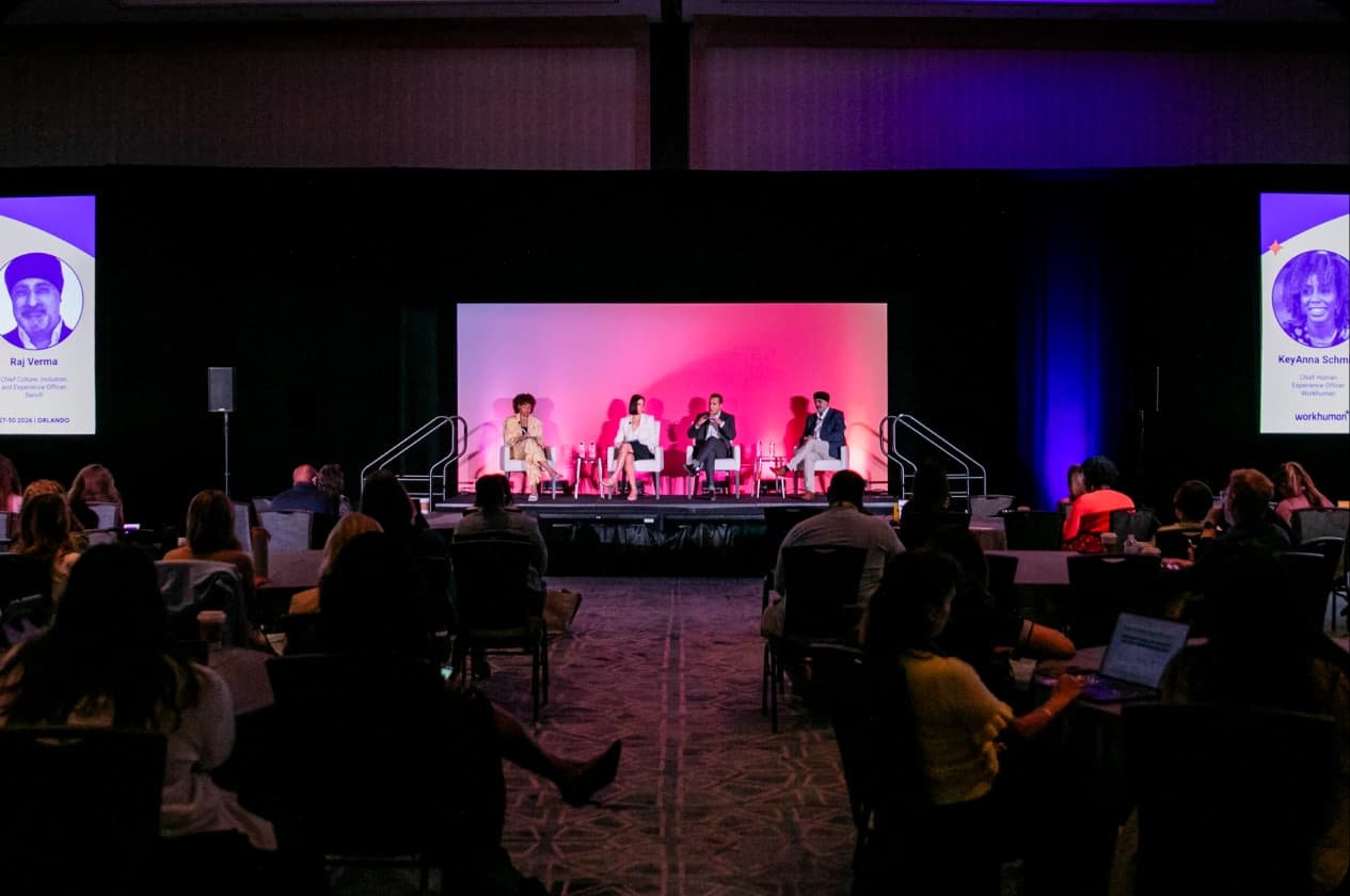 A panel discussion taking place on a stage with four speakers seated in front of a bright, gradient backdrop. The audience, visible in the foreground, consists of people seated at tables, engaged with their devices. Two names, Raj Verma and KeyAnna Schmid, are displayed on screens beside the speakers.