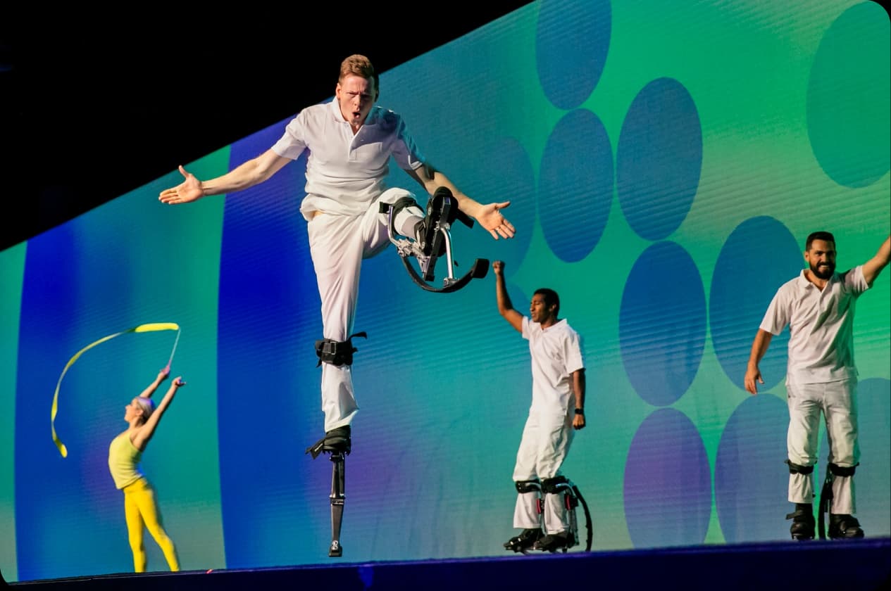 A performer in a white outfit is jumping with one leg raised, using spring-like leg apparatus, while two other individuals in similar attire stand beside him, one raising a fist in celebration. In the background, a colorful screen features large, abstract circles. Another performer in a yellow outfit twirls a ribbon.
