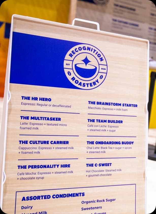 A wooden signboard displays a menu for a coffee roastery, titled “Recognition Roastery, Est 2026.” The menu lists various coffee drinks, including "The HR Hero" (Espresso: Regular or decaffeinated), "The Brainstorm Starter" (Macchiato: Espresso + milk foam), and "The C-Sweet" (Hot Chocolate: Steamed milk + gourmet chocolate). It also includes a section for assorted condiments like dairy milk and sweeteners.