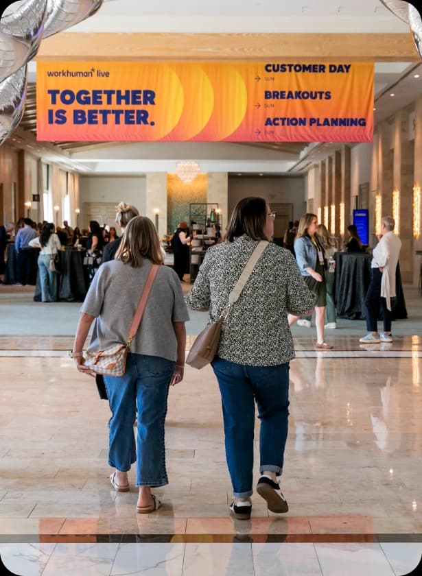 Two women are walking away, viewed from behind, in a spacious indoor event space. A large banner above them reads "workhuman live" and "TOGETHER IS BETTER," with additional text about "CUSTOMER DAY," "BREAKOUTS," and "ACTION PLANNING." In the background, several people are engaged in conversation around tall tables.