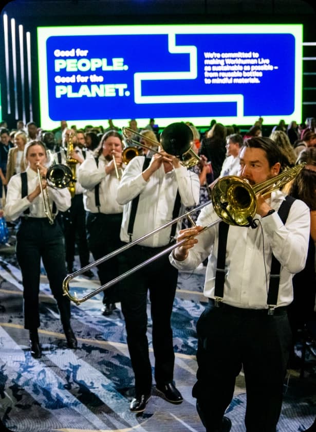 A group of musicians in formal attire plays trombones in a crowded venue, with a large illuminated sign in the background displaying the text "Good for PEOPLE. Good for the PLANET."