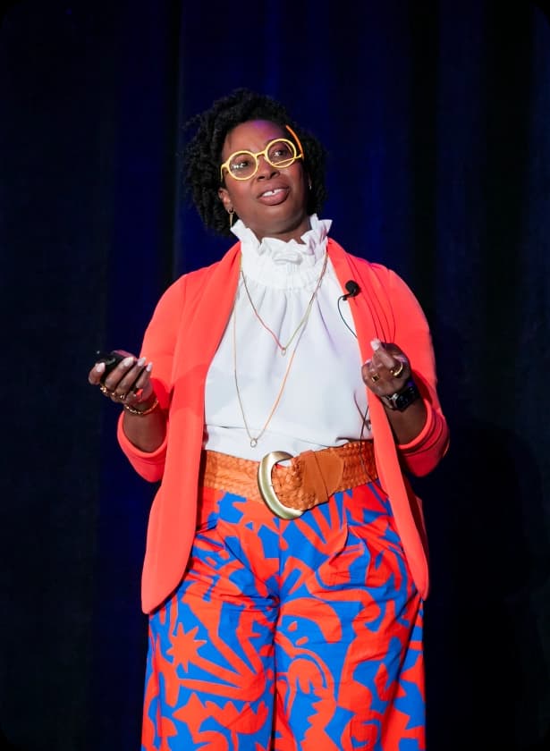 A person wearing large round glasses and an orange blazer stands on a stage, holding a small device in one hand. They are dressed in a white blouse and patterned blue and orange pants, with a large belt and necklace visible, and a microphone is attached to their clothing. The background is dark.