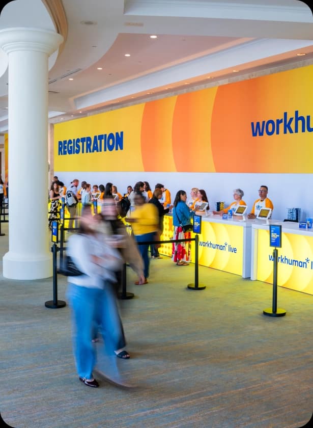A crowded registration area with a large wall displaying the word "REGISTRATION" in blue against a yellow background, and the text "workhuman live" below. Attendees are lined up at a reception desk where staff members in yellow shirts assist them, surrounded by a bright and open space.