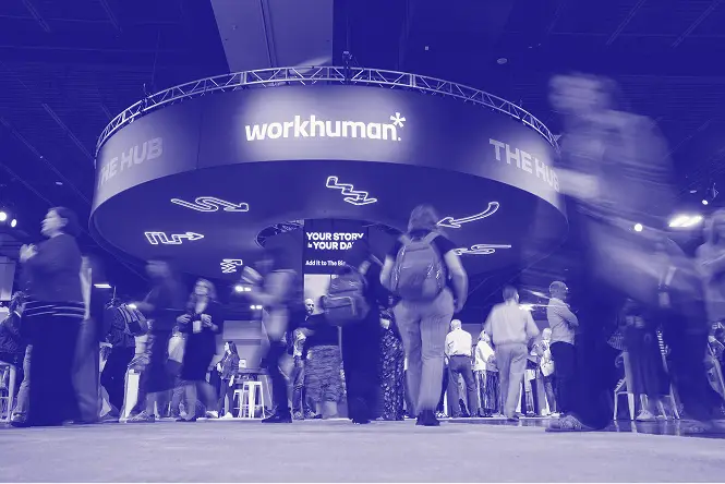A bustling Workhuman event with attendees mingling under a large overhead display that reads "workhuman" and "THE HUB." Additional text visible includes "YOUR STORY" and "YOUR DAY." The atmosphere is lively, capturing a moment of connection among participants.