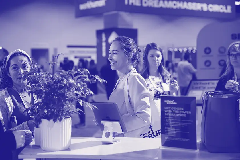 A smiling woman holding a tablet engages with others at a conference booth, where a small plant is placed on a table. The background features several attendees, and a sign reads, "LIFT OTHERS WITH THE POWER OF GRATITUDE." A second sign with the text "Dare to RUP" is partially visible. The setting conveys a lively atmosphere of connection and appreciation among participants.