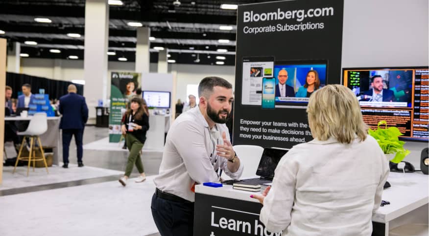 A business professional is engaged in conversation at a booth displaying Bloomberg's corporate subscriptions. The booth features a large sign with the text 'Bloomberg.com Corporate Subscriptions' and a graphic highlighting the benefits of their services. In the background, other attendees are interacting in the event space. Two monitors display news segments and stock information.