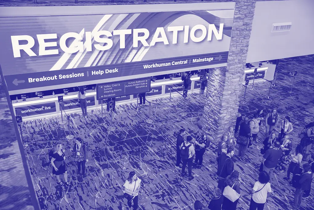 A bustling registration area at a Workhuman event, featuring a large "REGISTRATION" sign prominently displayed. Attendees are gathered in front of registration counters labeled for various breakout sessions and help desk services. The environment is lively with people interacting, and the floor features a stylized pattern, contributing to the event's vibrant atmosphere.