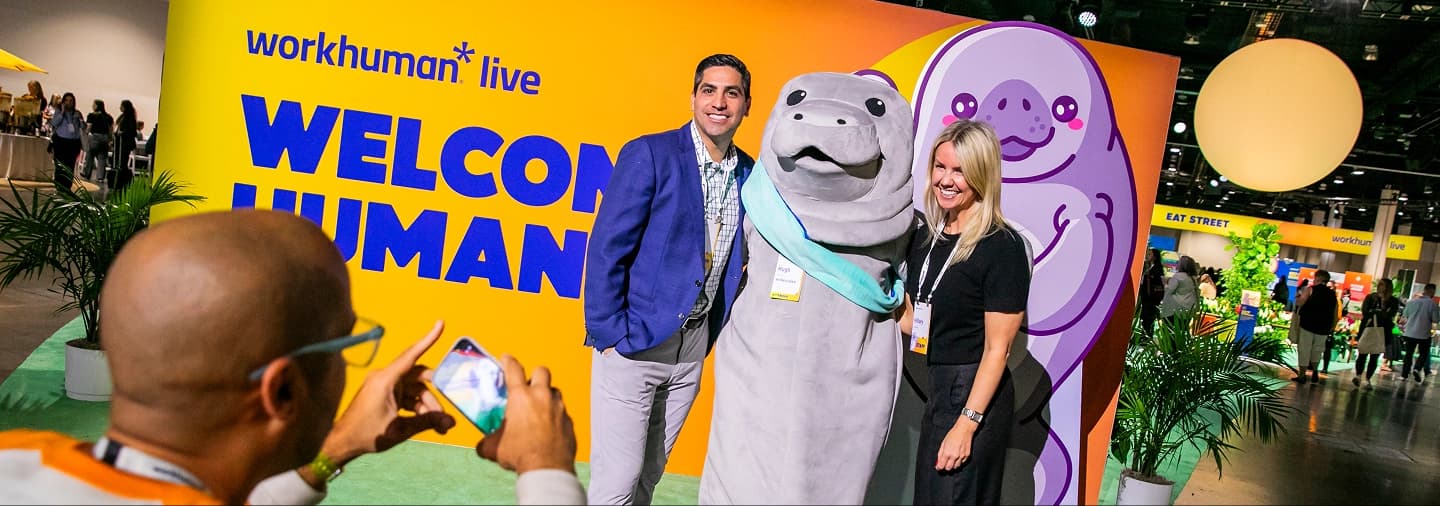 Attendees pose with the Workhuman mascot, Hugh, in front of a bright yellow and orange “Workhuman Live” welcome display while another person photographs them at the event.