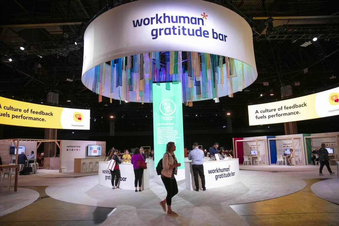 A vibrant Workhuman Gratitude Bar display at a conference features colorful fabric streamers hanging from a circular overhead sign. Attendees engage with the booth, which highlights messages about fostering a culture of feedback that enhances performance. Nearby, additional displays showcase the Workhuman cloud, while a digital screen emphasizes the importance of feedback in workplace conversations.
