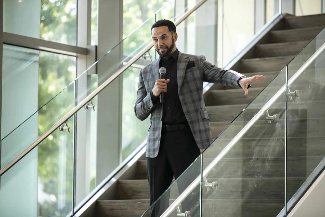 A man in a plaid blazer stands on a staircase, holding a microphone and gesturing with his hand as he speaks. The background features large glass windows with greenery visible outside, creating a bright and inviting atmosphere.