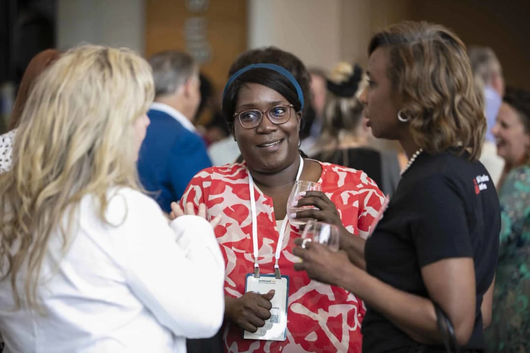 A group of attendees at a Workhuman event are engaged in conversation, with a focus on a woman wearing a red patterned top and glasses, holding a drink. She has a friendly expression and is interacting with two other women, one in a white jacket and the other in a black shirt. The background includes other event participants and a blurred setting, indicating a lively atmosphere. A badge can be seen around the woman's neck, indicating her participation in the event.