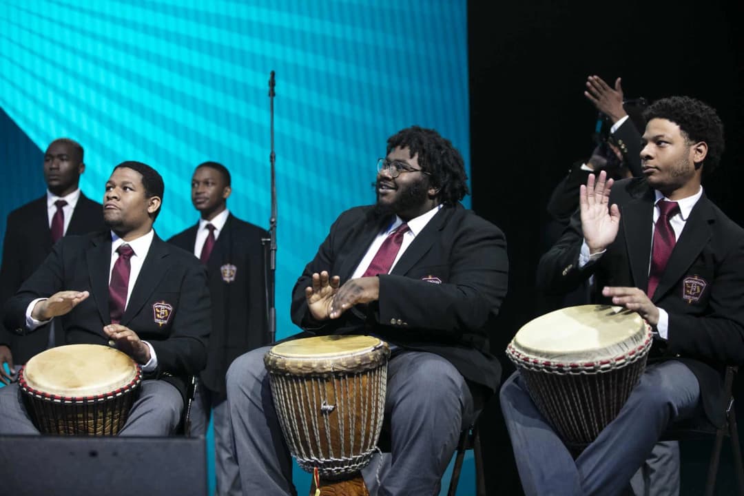 A group of four men performing on stage, seated and playing drums. They are dressed in black blazers with ties, while the background features a blue and turquoise lighting design. Some men in the background appear to be standing, and there is a microphone stand visible. The individuals are engaged in a rhythmic performance as they interact with each other and the crowd.