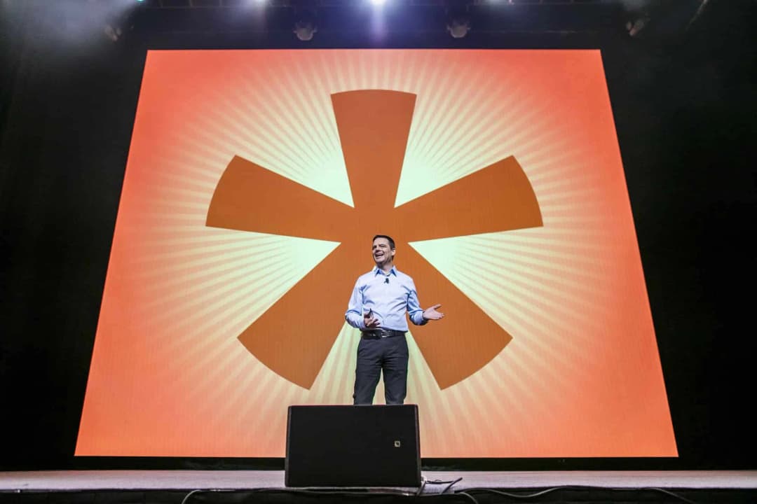 A speaker stands on stage before a large backdrop featuring a vibrant radial design with an asterisk symbol at its center. The speaker is smiling and gesturing, creating an engaging atmosphere. The overall setting conveys a sense of excitement and energy, likely part of a presentation or event.