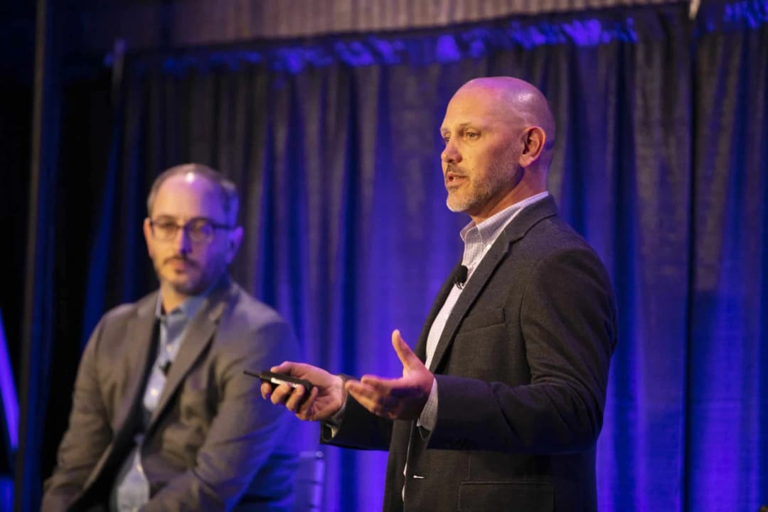 A presenter speaking on stage while holding a device in one hand, gesturing with the other, and wearing a dark blazer over a light-colored shirt. Another man in a gray suit sits in the background, listening attentively. The backdrop consists of dark curtains with purple lighting, creating a professional atmosphere for the presentation.