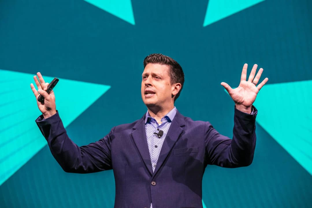 A person in business attire is speaking on stage, gesturing with both hands. He holds a microphone in his left hand and appears to be emphasizing a point. The background features large, vibrant geometric shapes in shades of teal and turquoise, contributing to a dynamic atmosphere typical of a conference setting.