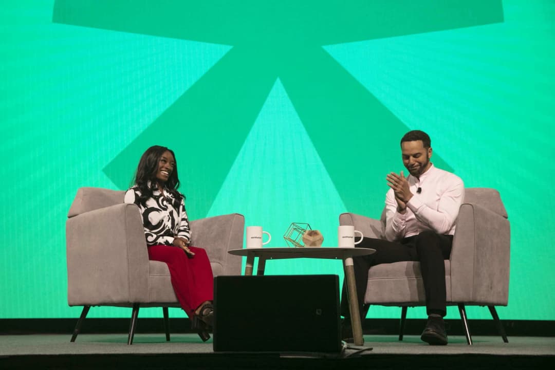 A woman and a man seated on stage in a conference setting, with the woman smiling brightly while the man claps his hands in appreciation. They are positioned in front of a vibrant green background featuring stylized patterns. The table between them has two coffee mugs and a decorative item.