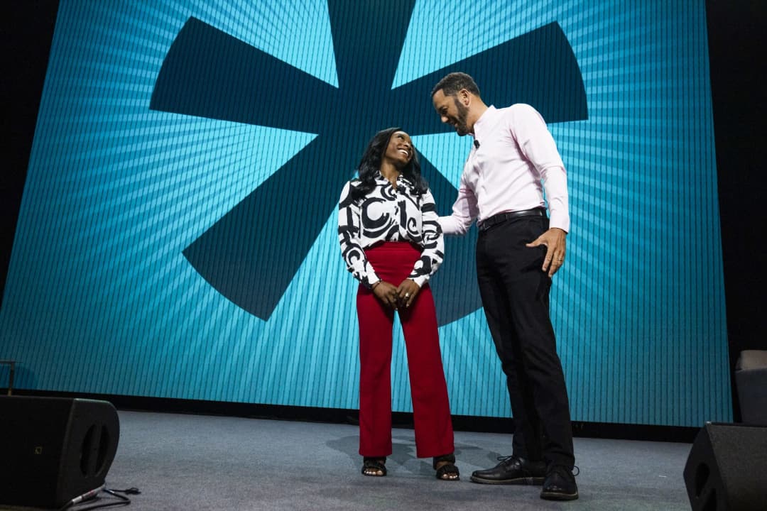 A woman wearing a black and white patterned blouse and red pants is smiling and standing on a stage, interacting with a man in a pink shirt and black pants. They are in front of a large backdrop featuring a stylized, large asterisk design. The scene captures a moment of connection and engagement between the two individuals during a presentation or event.