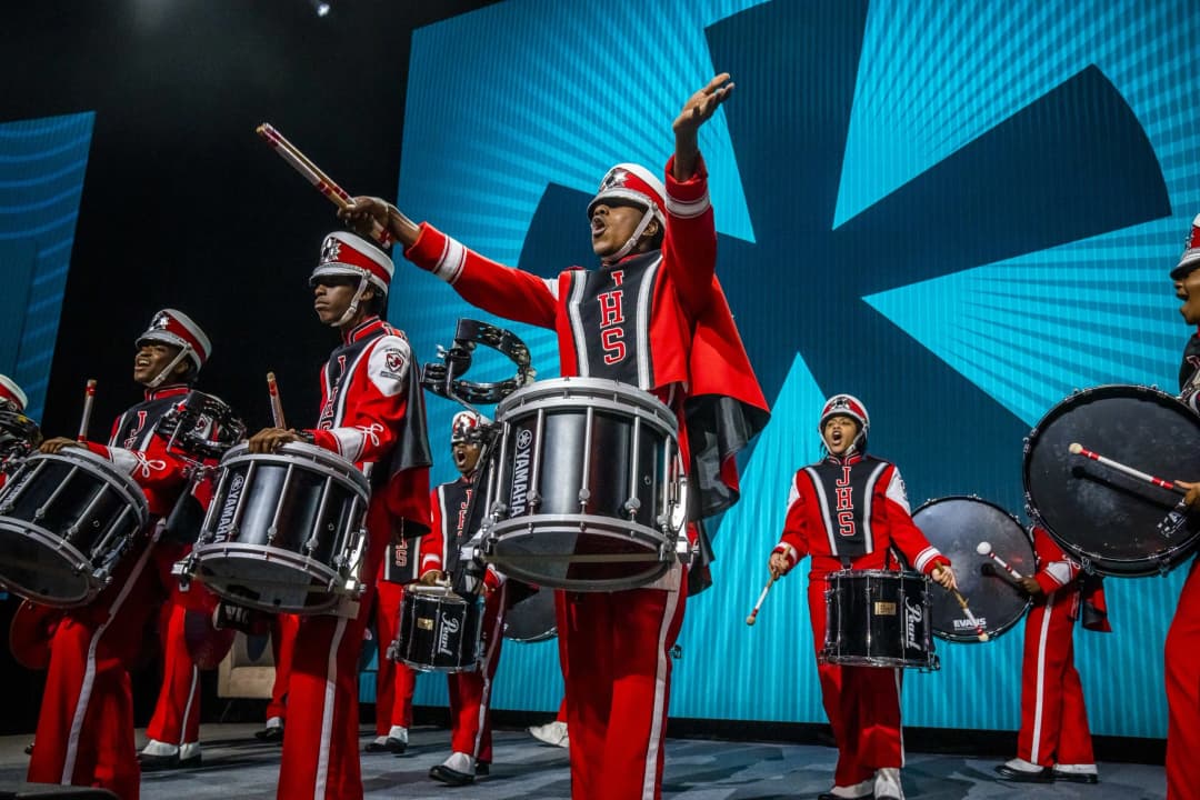 A group of drummers in vibrant red and black uniforms performs on stage, showcasing a lively and energetic display. One drummer in the center, wearing a hat, raises his arms energetically while others play their drums enthusiastically. The background features large, illuminated blue panels with a starburst design, adding to the festive atmosphere. The drums prominently display the brands 'YAMAHA' and 'Pearl.'